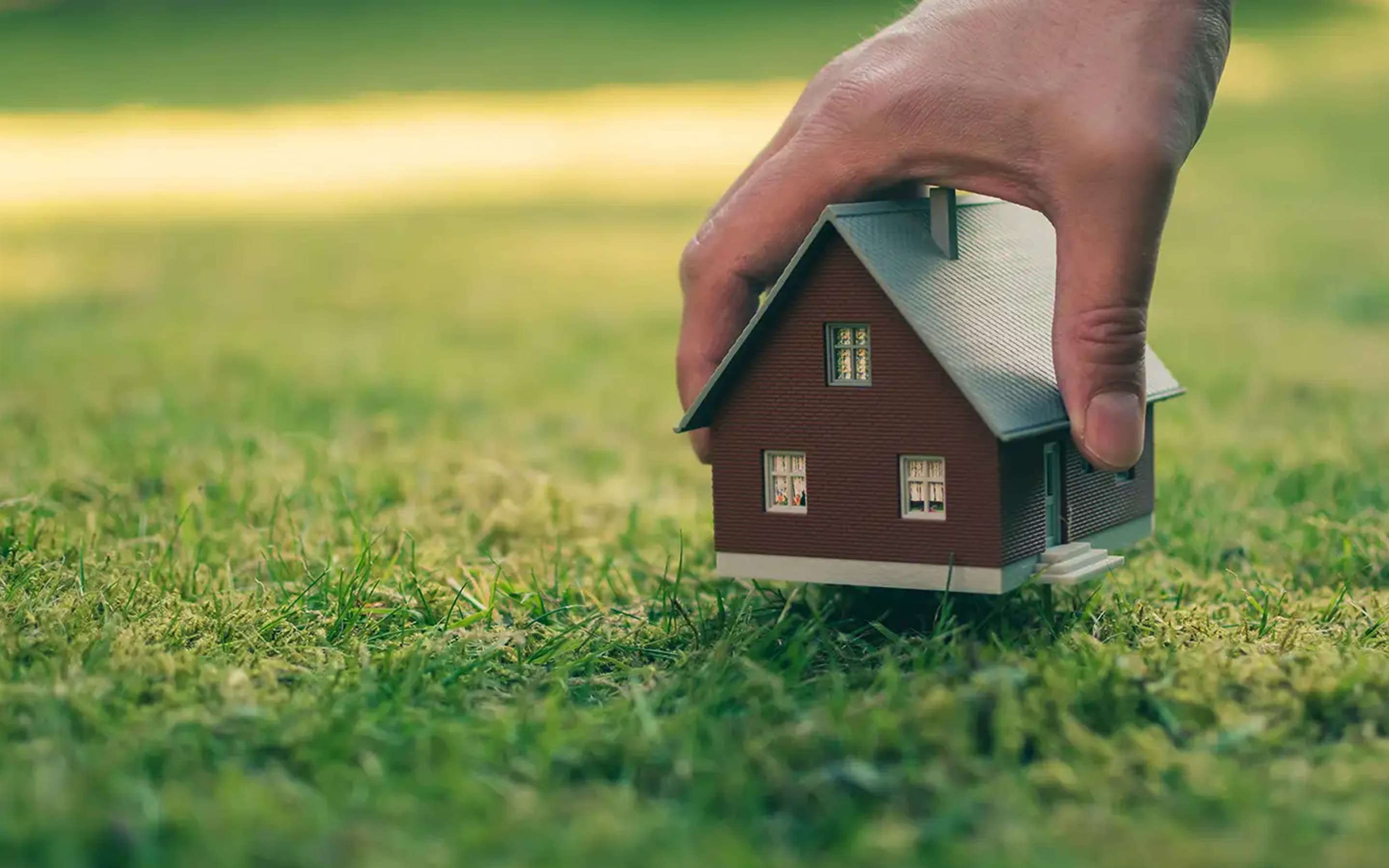 Hand greift ein Haus, welches auf einer Wiese steht