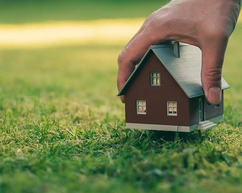 Hand greift ein Haus, welches auf einer Wiese steht